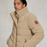 Woman wears a natural borg jacket with brown trim and brown leather patch on a grey background