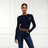Holland Cooper Princeton Crew Neck Knit Variant Colour Ink Navy.
A woman modeling a dark navy blue long-sleeve knit sweater with a small gold embroidered logo on the left chest, paired with dark blue jeans and a brown belt, standing against a white paneled wall.