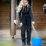 Woman wears a pair of black full chaps and black base layer whilst holding a hose on a yard setting