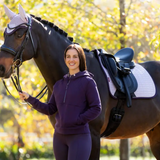 Woman in a purple outfit standing next to a horse with a saddle in an outdoor setting.