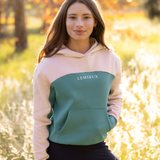 LeMieux AW26 Young Rider Sadie Hoodie Variant Colour Aloe.
A model wearing a colorblock hoodie with a pale pink hood and sleeves, a aloe colour body panel, and thin cream piping details. The brand name 'LEMIEUX' is printed in white across the chest. on faded meadow background.