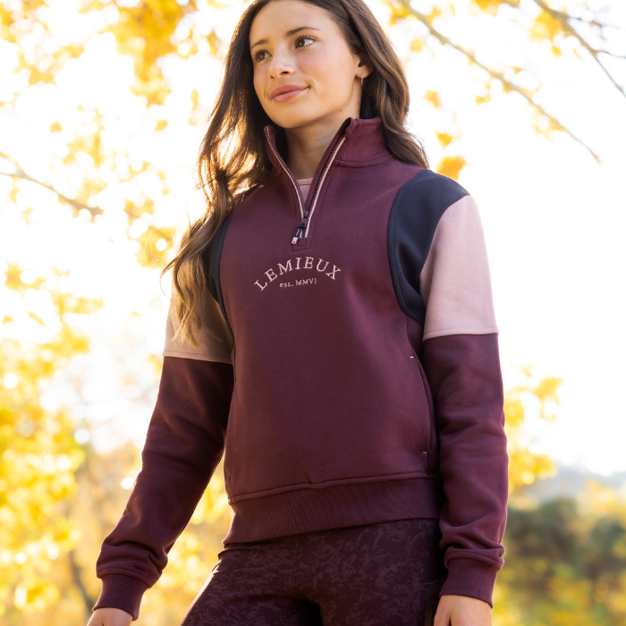 LeMieux AW26 Young Rider Viola Quarter Zip Sweat Variant Colour Bramble.
A woman wearing a color-blocked quarter-zip sweatshirt by Lemieux in bramble, navy, and light pink, standing outdoors against a bright, sunlit background of yellow autumn leaves.