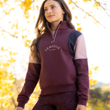 LeMieux AW26 Young Rider Viola Quarter Zip Sweat Variant Colour Bramble.
A woman wearing a color-blocked quarter-zip sweatshirt by Lemieux in bramble, navy, and light pink, standing outdoors against a bright, sunlit background of yellow autumn leaves.