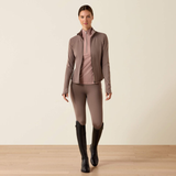 Woman wears a brown full zip jacket, riding leggings and black riding boots against a beige background