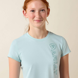 Girl wears pale blue t-shirt with ARIAT logo on a beige background