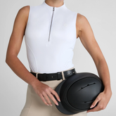 Person wears a white sleeveless base layer holding a black riding hat on a grey background
