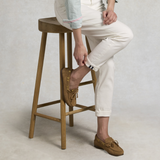 Person sitting on a wooden stool wearing white trousers and brown loafers.
