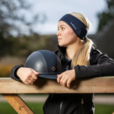 LeMieux AW26 Ear Warmer Headband Variant Colour Indigo. A model with blond hair wearing a blue headband, holding a riding helmet leaning against a gate on faded garden background.