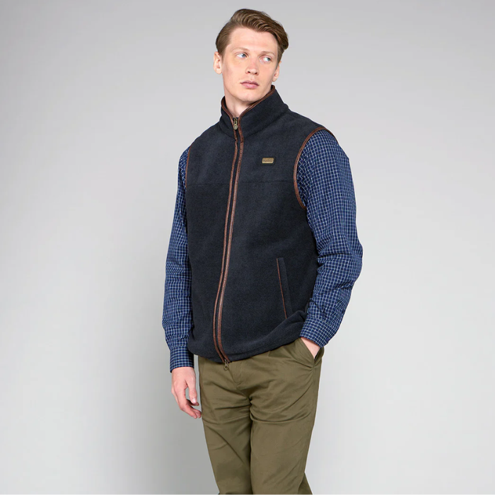 Man wearing a navy gilet over a blue checkered shirt with green pants on a grey background