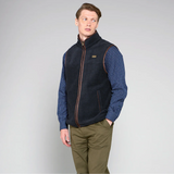 Man wearing a navy gilet over a blue checkered shirt with green pants on a grey background