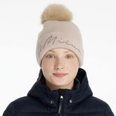 Person wearing a beige knit beanie with a fur pom-pom and a navy jacket on a white background