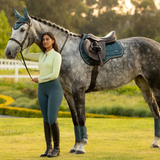 Woman in equestrian attire standing next to a grey horse in a field