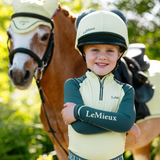 Child wearing a LeMieux helmet and riding jacket standing next to a horse in an outdoor setting.
