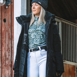 Woman wearing a floral base layer, white riding breeches and a black coat stands near a stable