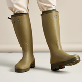 Person wears a pair of green wellington boots and cream trousers against a neutral background