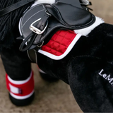 Horse wearing a black saddle with a red pad and white trim, standing on a concrete surface.