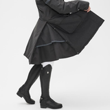 Person wears a black coat and long black riding boots against a white background