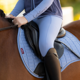 Rider sits in a saddle with a blue saddle pad on a brown horse and a blurred outdoor background