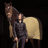 Woman in a black jacket standing next to a horse with a beige rug on a dark background