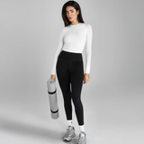 Woman in white long-sleeve top and black leggings holding a yoga mat on a grey background