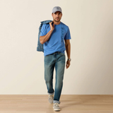 Man wearing a blue t-shirt, jeans, and cap holding a denim jacket against a beige wall.