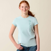 Girl wears pale blue t-shirt with ARIAT logo on a beige background