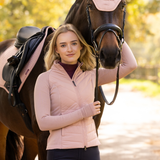 LeMieux AW26 Maddie Hybrid Jacket Varient Wild Rose. A woman stands beside a dark brown horse outdoors, wearing a light pink ribbed zip-front equestrian jacket over a dark top. The horse has a pink and black saddle with a bridle, and the background is a softly blurred outdoor path and trees.