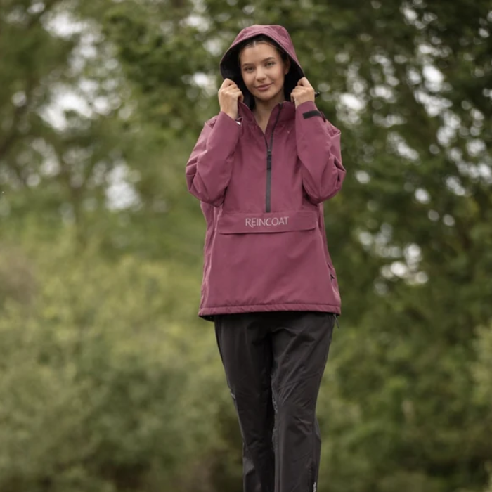 Reincoat Smock Lite Varient Bloom. Woman wearing a Bloom coloured jacket with a black zip and kangaroo style pocket with "REINCOAT" branding on a blurred forest background. 