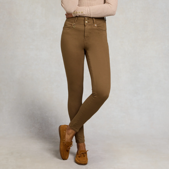 Holland Cooper Jodhpur Jean Variant Chesnut. A model from the waist down wearing slim-fit brown skinny jeans, a beige ribbed sweater, and brown suede boat shoes, standing against a textured light gray studio backdrop.