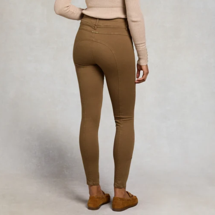 Holland Cooper Jodhpur Jean Variant Chesnut. A person shown from the back wearing fitted ankle-length jeans in a warm tan/camel color, paired with a light beige long-sleeve top and brown suede flats, against a light gray studio background.