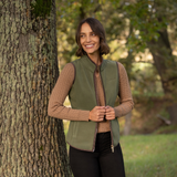 LeMieux AW26 Frankie Fleece Gilet Variant Colour Willow Green.
A woman stands outdoors next to a large tree trunk, wearing an willow green zip-front vest over a beige cable-knit sweater and black pants.