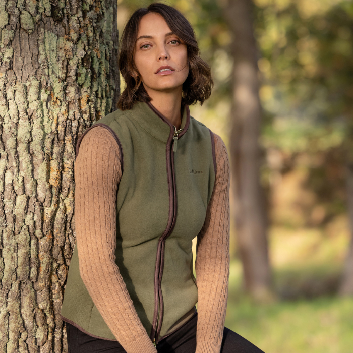 LeMieux AW26 Frankie Fleece Gilet Variant Colour Willow Green.
A woman stands outdoors next to a large tree trunk, wearing an willow green zip-front vest over a beige cable-knit sweater and black pants.