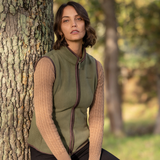 LeMieux AW26 Frankie Fleece Gilet Variant Colour Willow Green.
A woman stands outdoors next to a large tree trunk, wearing an willow green zip-front vest over a beige cable-knit sweater and black pants.