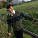LeMieux AW26 Mens Rugby Shirt Varient Olive. A man wearing a Lemieux long-sleeve rugby shirt in olive green and navy blue with a white collar, standing outdoors in a grassy field next to a wooden fence.