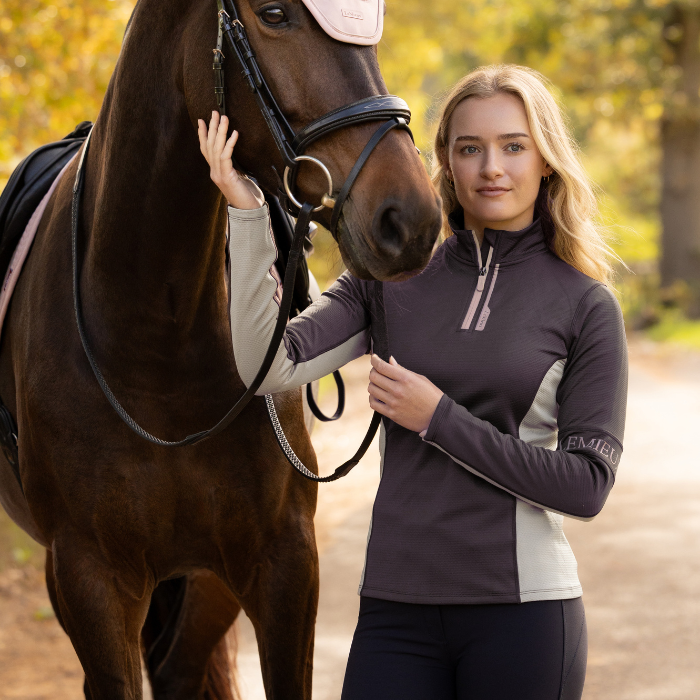 LeMieux AW26 Matilda Mid Layer Variant Colour Cocoa.
A woman stands outdoors beside a brown horse, wearing a fitted long-sleeve equestrian quarter-zip top with light beige side panels and a small zipper accent. The horse wears a bridle, and the background shows a softly blurred outdoor path and trees.
