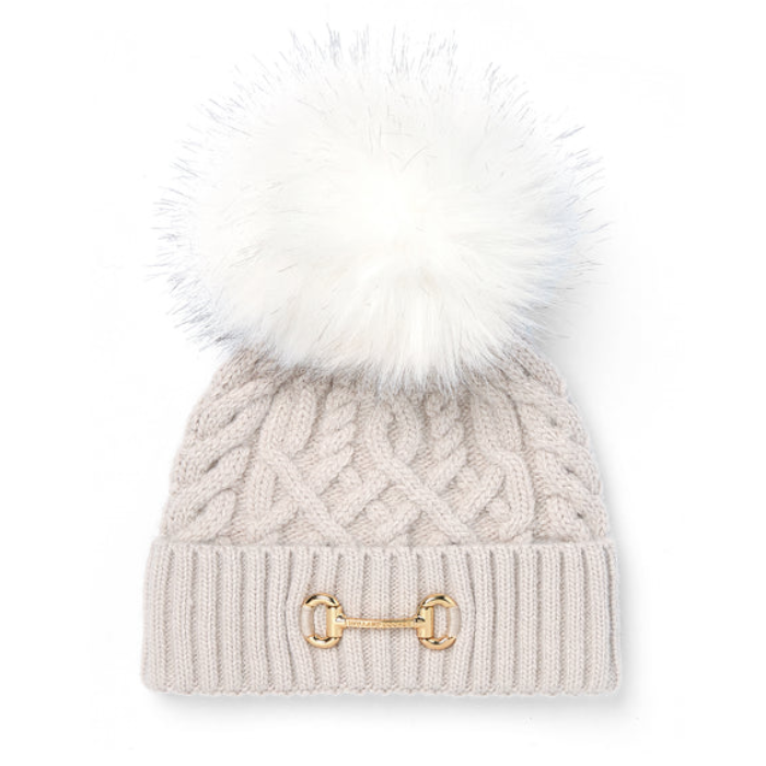 Holland Cooper Horsebit Bobble Hat Variant Taupe. A light beige cable-knit beanie with a large white faux-fur pom-pom and a gold horsebit detail on the front cuff, shown against a plain white background.