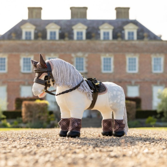 LeMieux AW26 Toy Pony Heritage Pad Varient Cocoa. A small white horse figurine or toy with a gray mane, black harness, and brown plaid leg wraps, standing on a gravel surface with a large brick building blurred in the background.