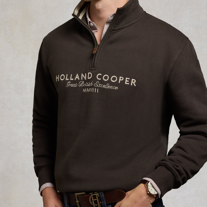 Holland Cooper Heritage Quarter Zip Sweat (Men's) Variant Bournvile. Man wearing a brown 1/4 zip sweatshirt with Holland Cooper branding across the chest in white agaisnt a grey background. 