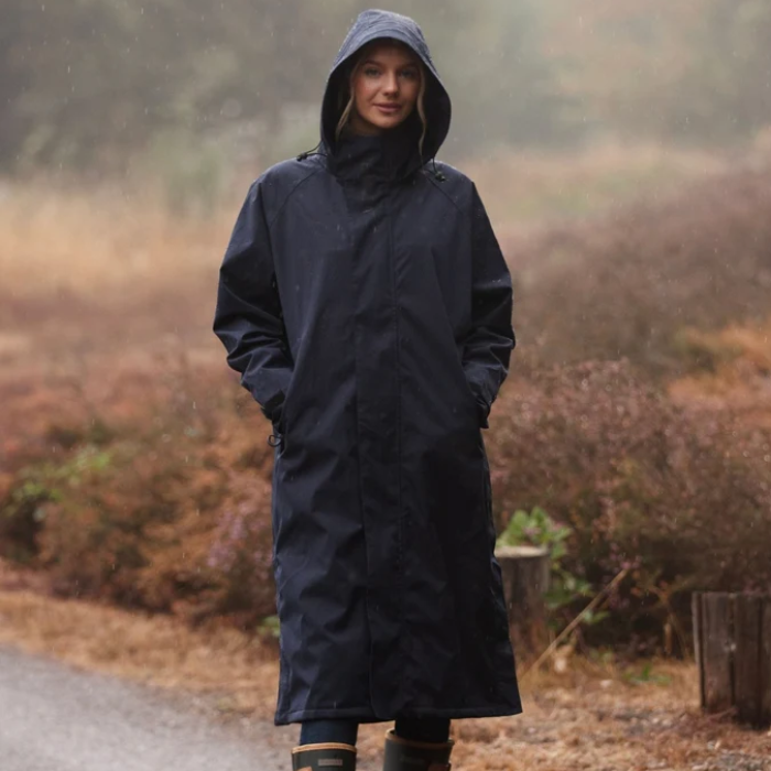A woman wearing a long, dark navy hooded raincoat stands on a path in the rain, with a blurred outdoor landscape of brown and green foliage in the background.