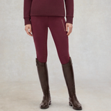 A model wearing tight equestrian riding breeches in deep burgundy, paired with a matching long-sleeve top and dark brown riding boots, standing against a neutral studio backdrop.