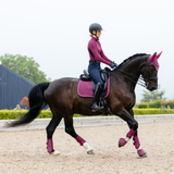 LeMieux AW26 Suede Dressage Square Varient Blackberry. A dark brown horse with a rider in equestrian attire, wearing matching magenta protective gear including a saddle pad, leg wraps, and ear covers, in an outdoor riding arena.