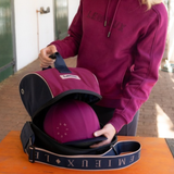 LeMieux AW26 Hat Bag Varient Blackberry. A person wearing a burgundy hoodie packs a matching burgundy and navy equestrian helmet into a zippered bag with a wide branded strap, standing outdoors at a table.