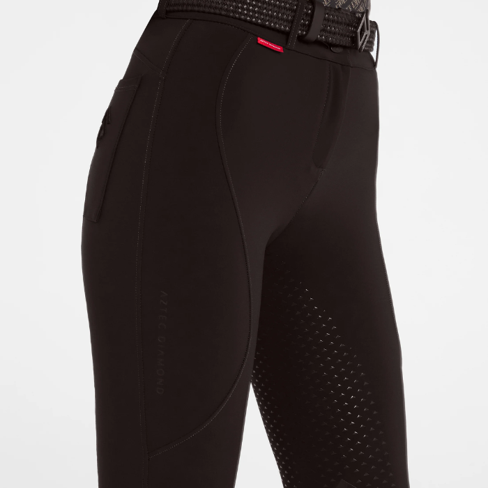 Close-up of brown equestrian riding breeches with textured grip panels on the seat and upper thigh, a small red brand tab, and a matching black patterned belt, shown against a light grey studio background.