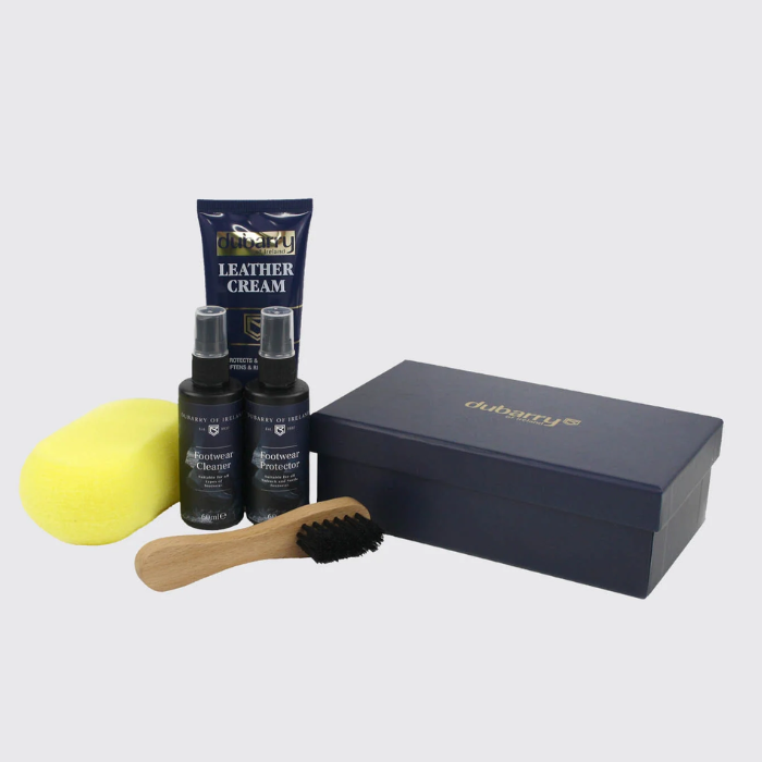 Dubarry leather care kit with a navy blue leather cream tube, two leather spray bottles, a yellow sponge, a wooden-handled brush, and a navy blue presentation box on a light gray background.