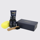 Dubarry leather care kit with a navy blue leather cream tube, two leather spray bottles, a yellow sponge, a wooden-handled brush, and a navy blue presentation box on a light gray background.