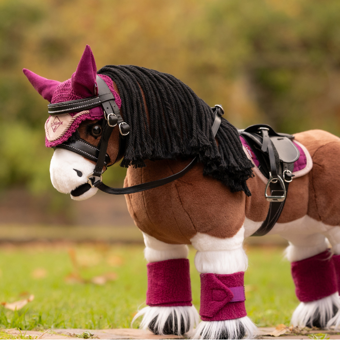 LeMieux AW26 Toy Pony Fly Hood Variant Colour Blackberry.
A plush toy horse wearing a blackberry colour equestrian costume, including a horned bridle, black faux-fur mane, saddle, and leg wraps, standing on grass with a blurred outdoor background.