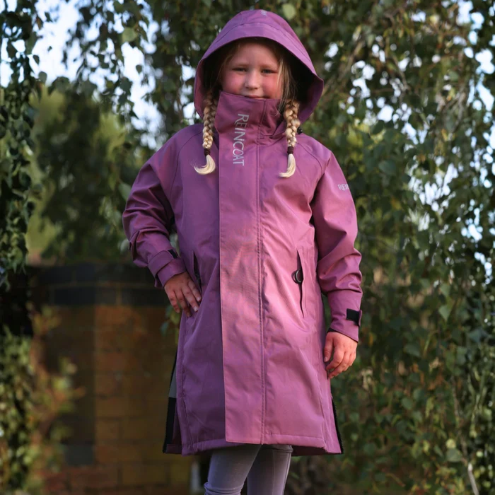 A young girl wearing a purple hooded raincoat with a high collar and drawstrings, standing outdoors in front of a blurred background of trees.