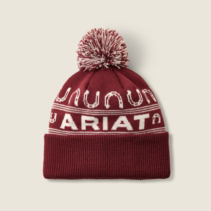 Ariat Ladies Apres Ski Beanie Varient Burgundy. Red and White beanie with large Ariat branding and horse shoe detailing with a red and white pom pom on a light background. 