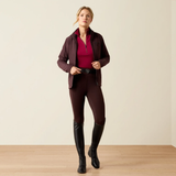 Ariat Women's Venture 2.0 Thermal Full Seat Tight Variant Chocolate Plum. A woman modeling equestrian apparel in a studio setting, wearing a dark burgundy zip-front jacket over a bright magenta top, matching dark burgundy riding breeches, and tall black riding boots against a plain beige wall and light wood floor.