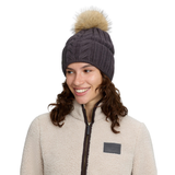 A woman wearing a dark gray cable-knit beanie with a beige faux-fur pom-pom, paired with a cream fleece jacket, against a solid white background.